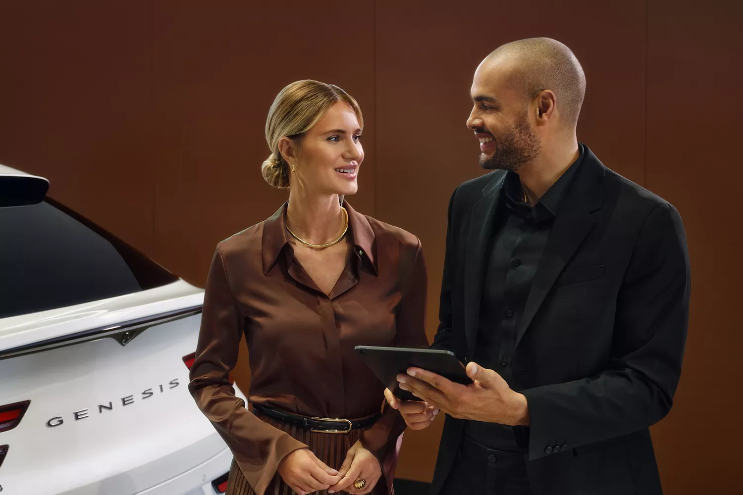 Two people wearing business casual clothes, a woman in brown and a man in black, smile while looking at a tablet.